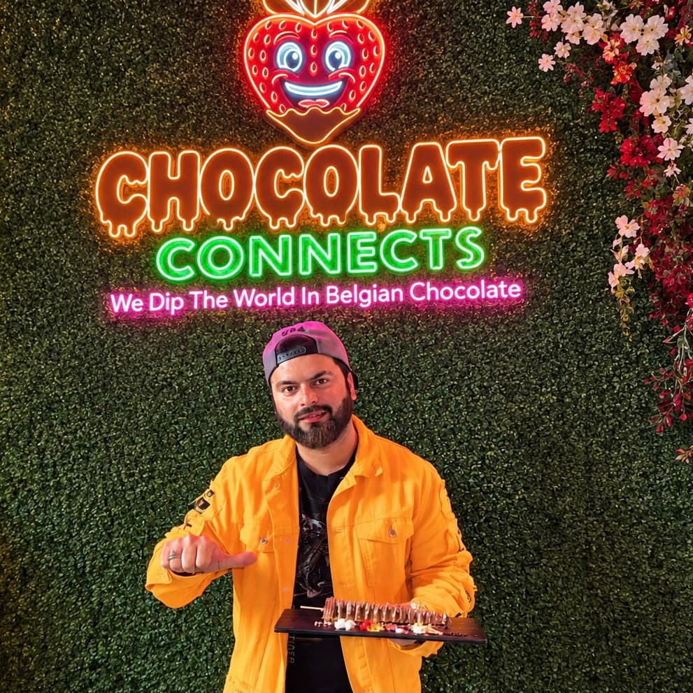 Muhammad Faisal Ishaq, founder of Chocolate Connects, holding a chocolate-dipped treat in front of the store neon